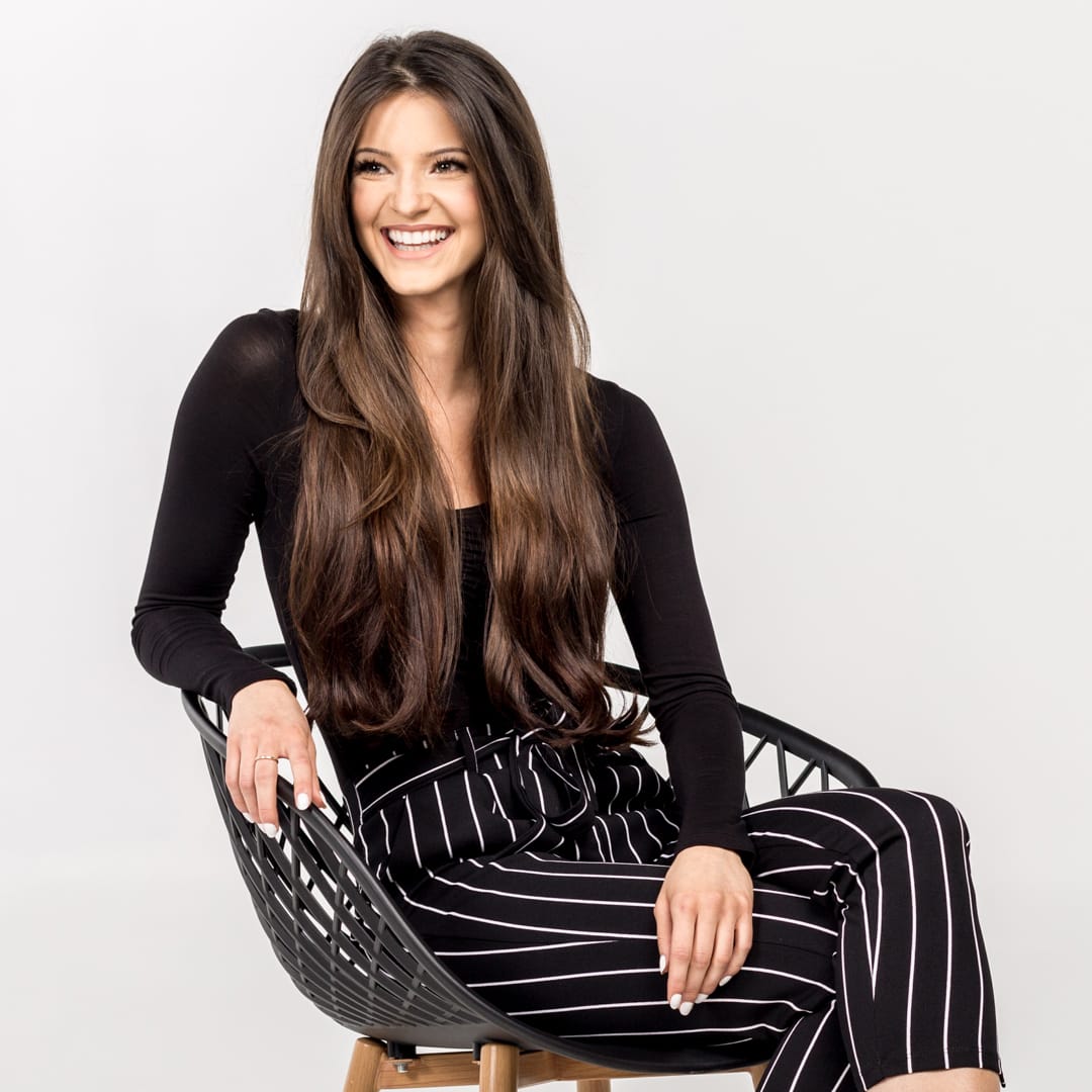 A woman with long brown hair sits on a black woven chair, smiling. She wears a black long-sleeved top and black-and-white striped pants.