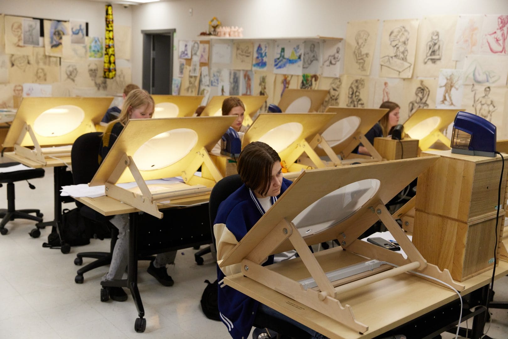 Students working at drawing desks in an art classroom with sketches displayed on the walls.