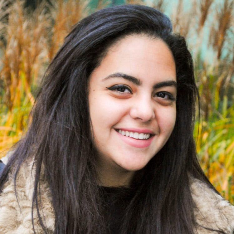 A person with long dark hair is smiling outdoors in front of tall grass and a blurred green background. CAT Alumni Nada Almessery appears vibrant and at ease in nature's embrace.