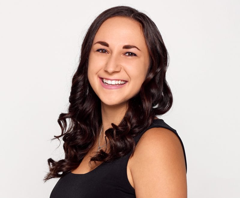 Ashlee Bardos, smiling with long, wavy, dark hair, wearing a sleeveless black top, against a plain white background.