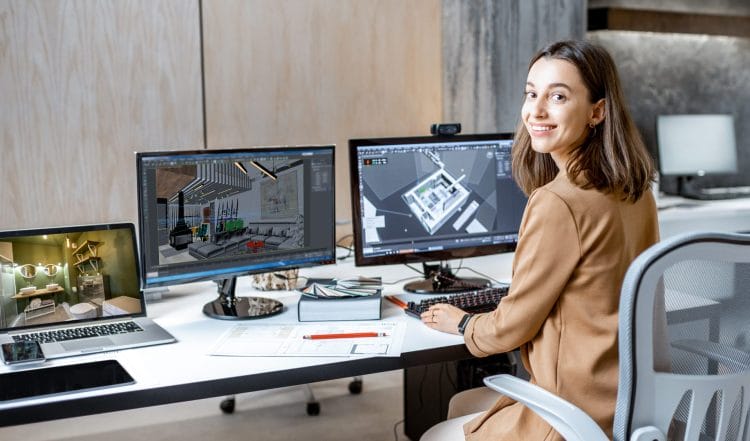 Dynamic photo featuring an interior design student enthusiastically working on a computer, surrounded by three monitors displaying different rooms in a house and a rendering of a house. The student faces the camera with a smile, exuding passion and dedication within the Centre for Arts and Design's innovative environment.