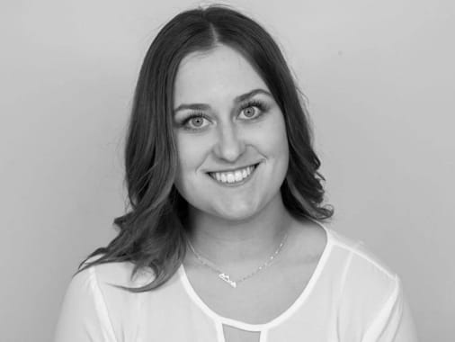 Black and white portrait of a woman, Taylor Woolf, with long, wavy hair smiling at the camera, wearing a light-colored blouse and a necklace.