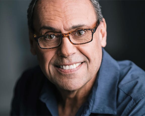 Damon Gregory, smiles at the camera and is wearing glasses and a blue shirt looking at the camera.