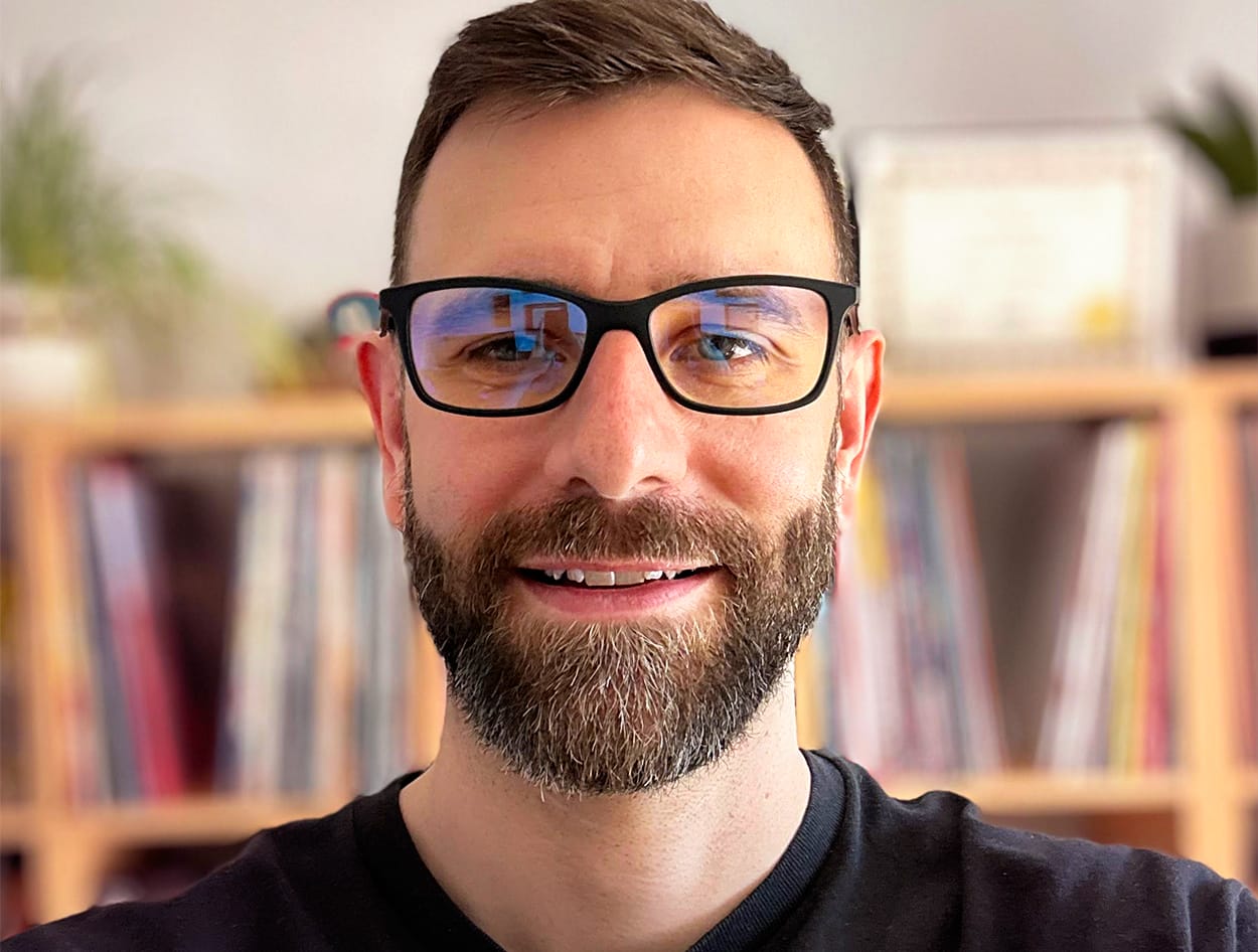 David Fenoulhet with glasses and a beard smiles at the camera in a room with a blurred bookshelf background.