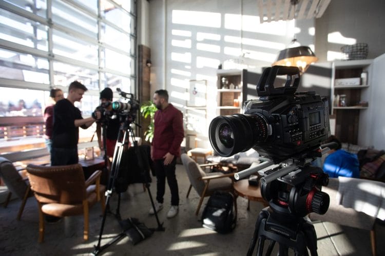 A group of people setting up video cameras and equipment in a well-lit room with large windows, chairs, and a table.