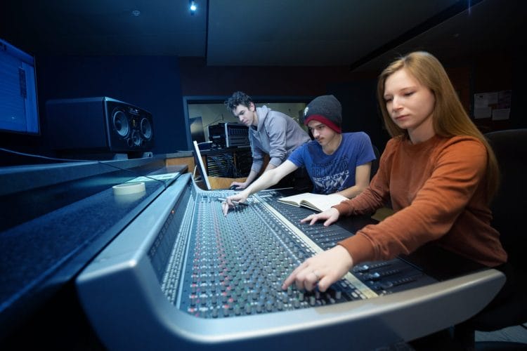 Three students are working on a large audio mixing console in a recording studio at Centre for Art and Design in Kelowna. The person on the left is adjusting controls, the person in the middle is taking notes, and the person on the right is operating the console.