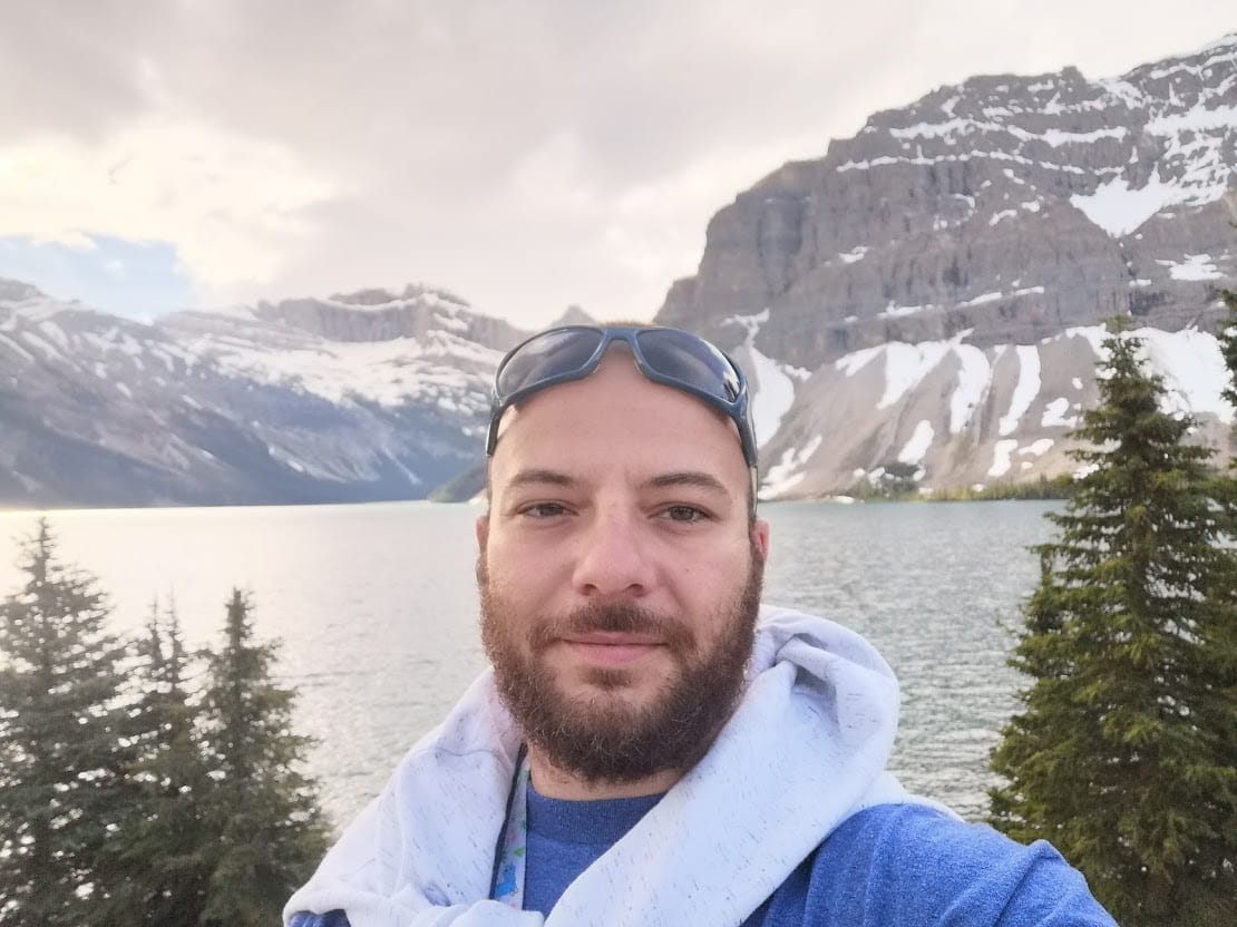 A man with a beard taking a selfie in front of a lake.