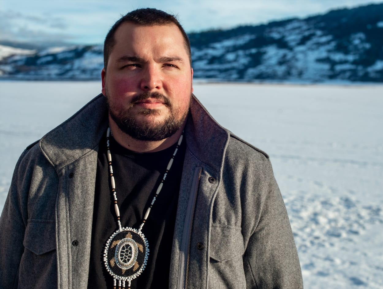 A man with a beard stands in a snowy landscape, wearing a gray coat and beaded necklace, looking towards the distance. Snow-covered mountains are visible in the background under a blue sky. This headshot captures CAT Alumni Jay Falkus in his element, exuding quiet strength and introspection.