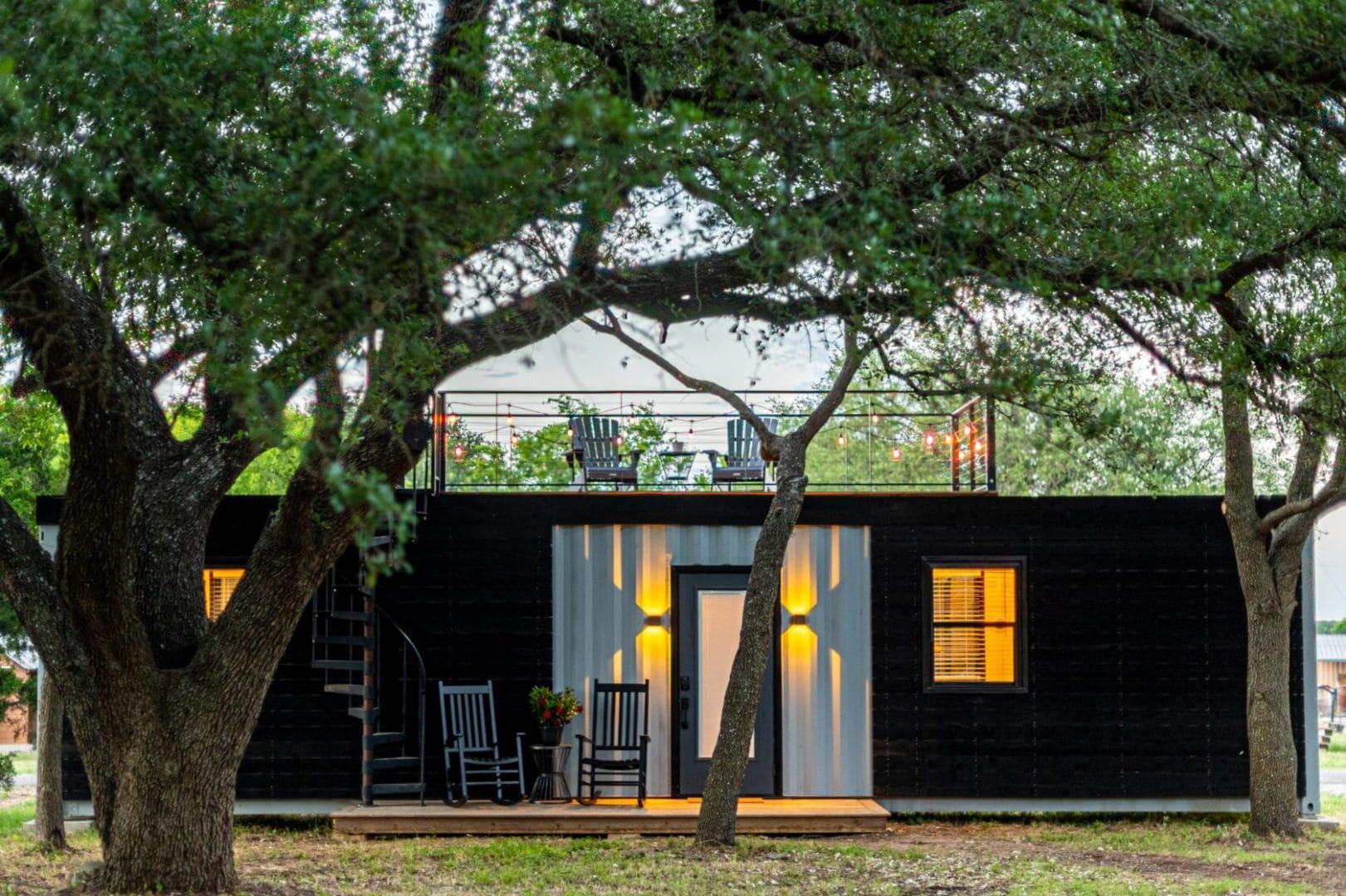 A black container house with a deck in the woods.