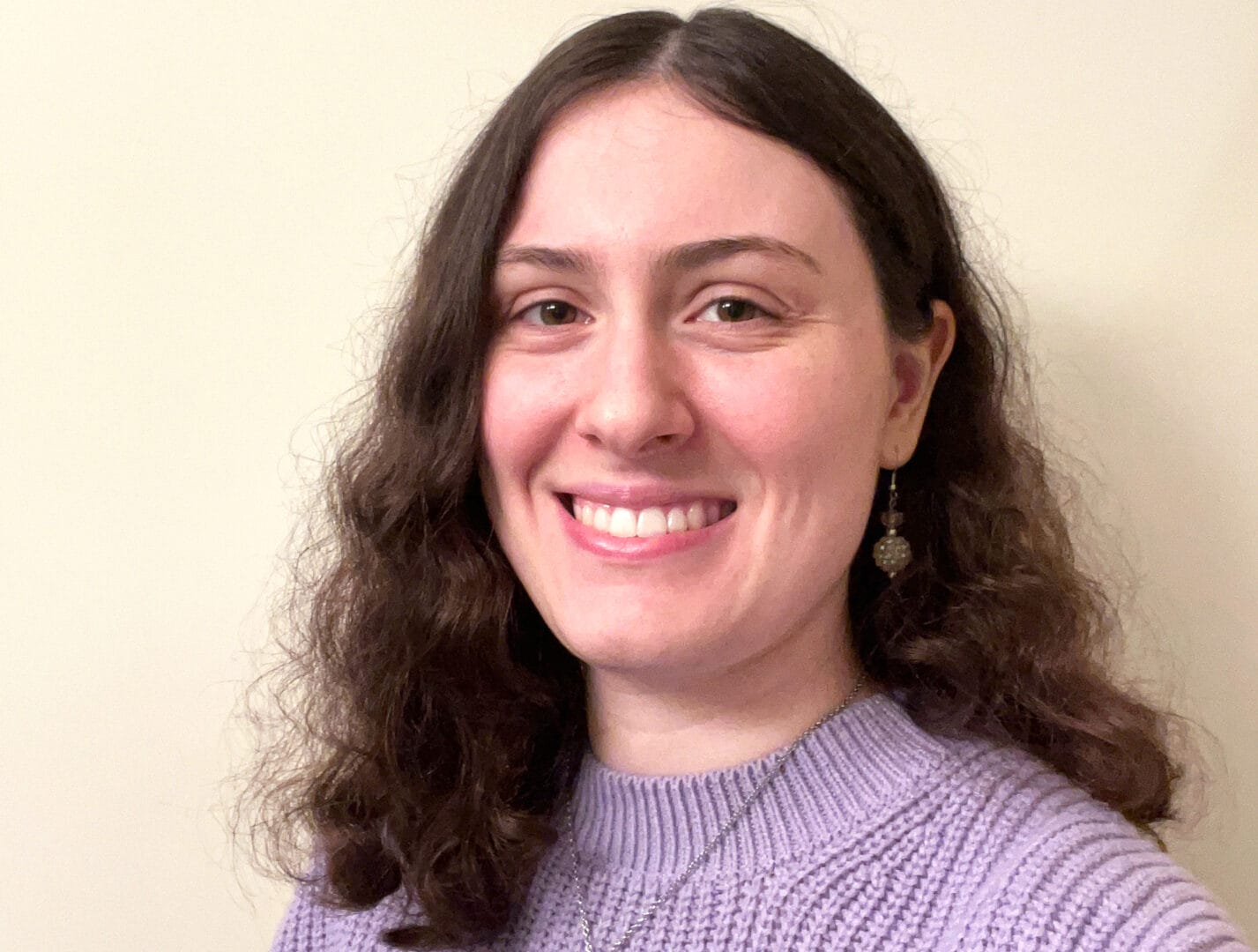 A person with shoulder-length wavy hair, wearing a purple sweater and drop earrings, smiles at the camera against a plain background.