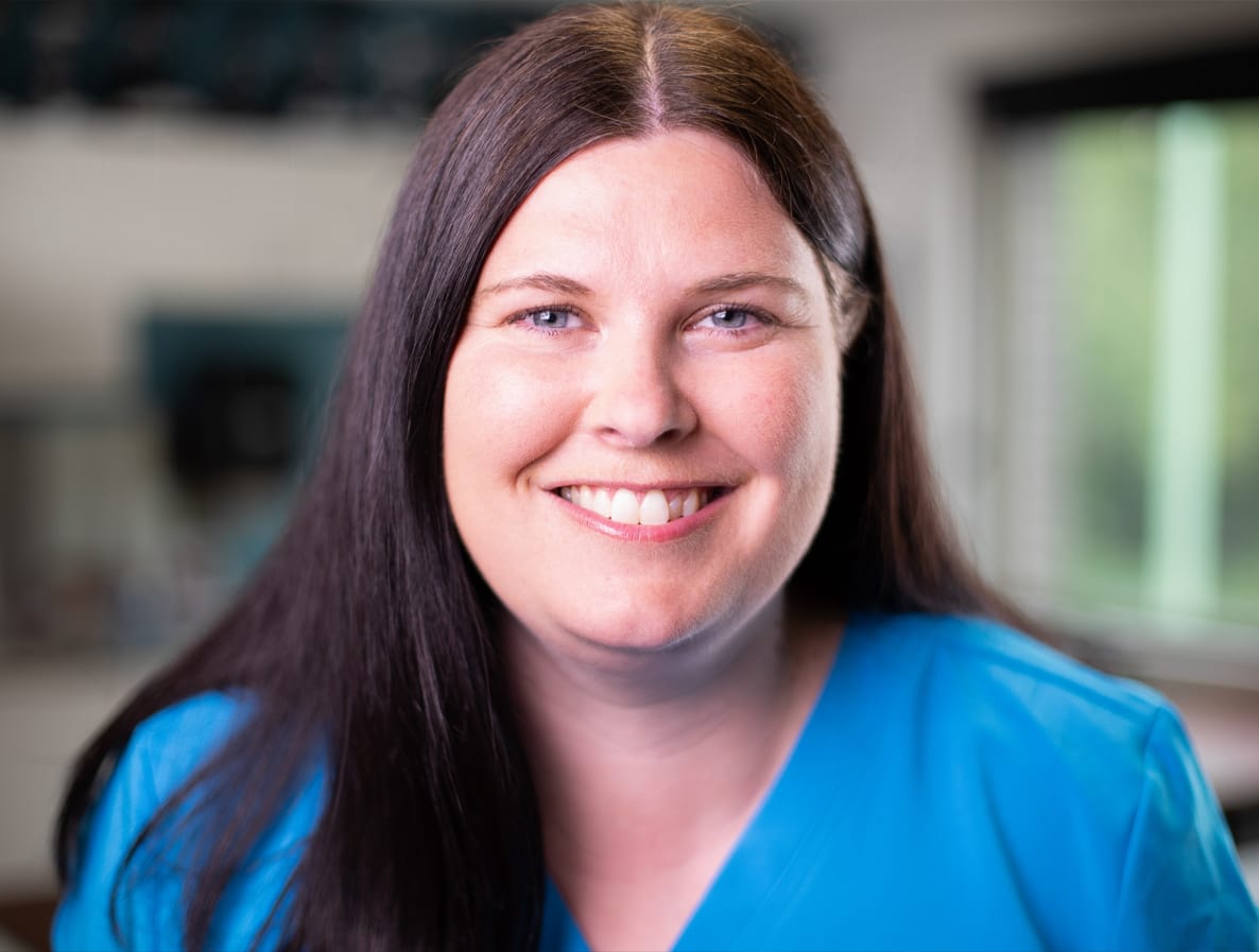 A woman with long dark hair smiles while wearing a light blue top in a brightly lit room.