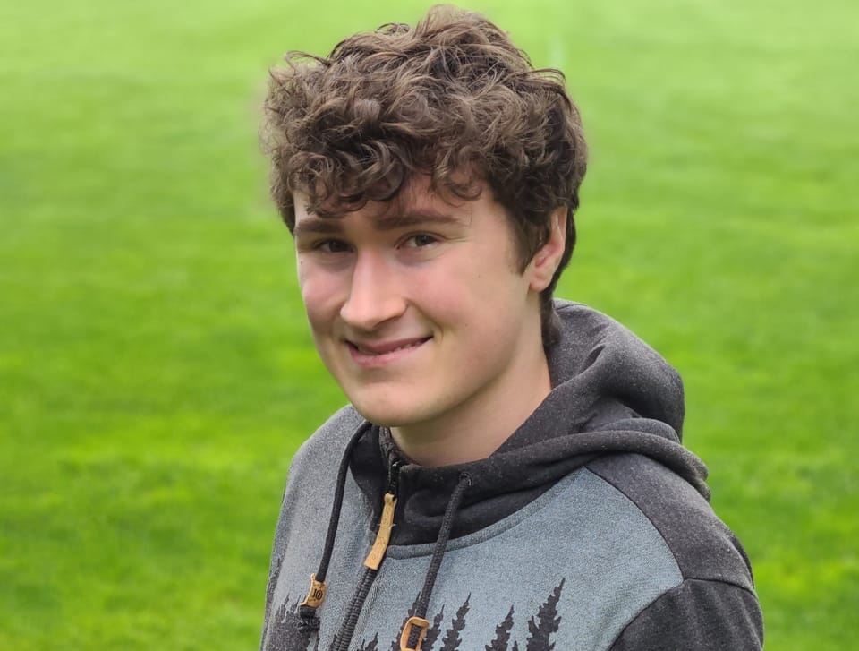 A person with curly hair wearing a hoodie stands on green grass, smiling at the camera.