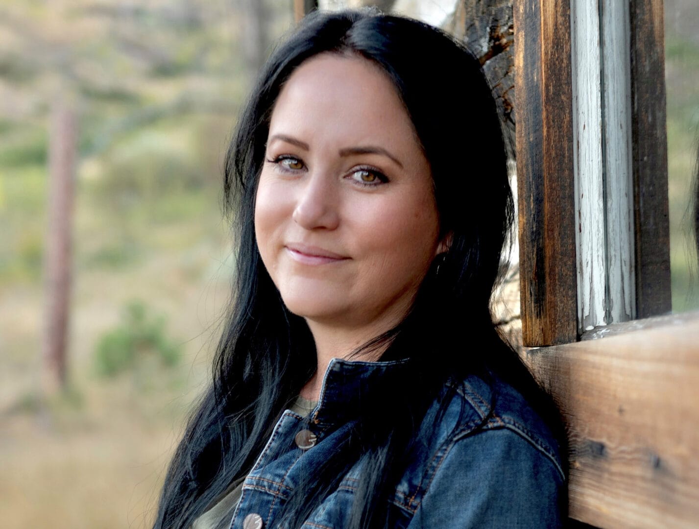 A woman with long dark hair and a denim jacket leans against a rustic wooden wall outdoors, smiling softly at the camera.