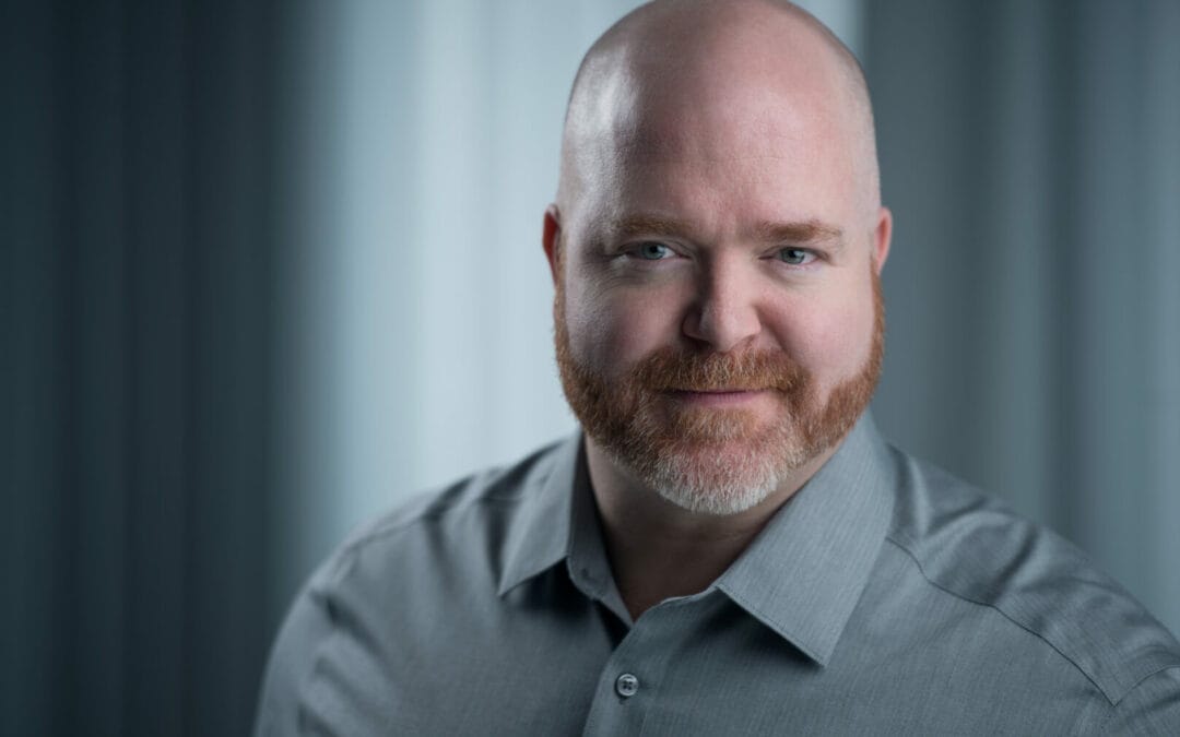 Peter Church, a man with a red beard and mustache, wearing a gray shirt, looks directly at the camera. The background is softly out of focus.