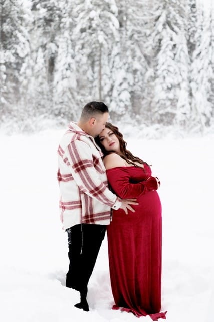 A couple embracing in a snow-covered forest, with the man kissing the woman's forehead and the woman in a red dress caressing her pregnant belly.