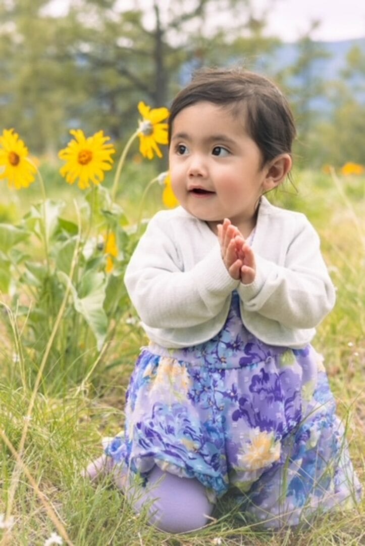 Toddler girl in a floral skirt and white sweater clapping hands among yellow flowers in a grassy field.