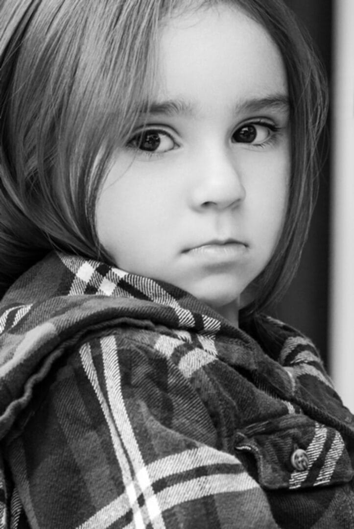 Close-up of a young girl in black and white, wearing a plaid shirt, looking thoughtfully towards the camera.