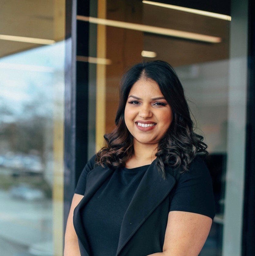 A smiling woman in a black suit standing in front of a window.