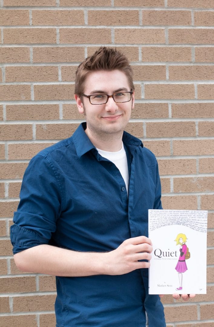 A young man holding a book in front of a brick wall.