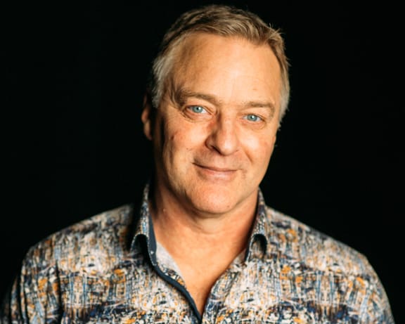 Roark Critchlow, instructor at CAT, showing short light hair and blue eyes is smiling slightly. He is wearing a patterned collared shirt and is set against a dark background.