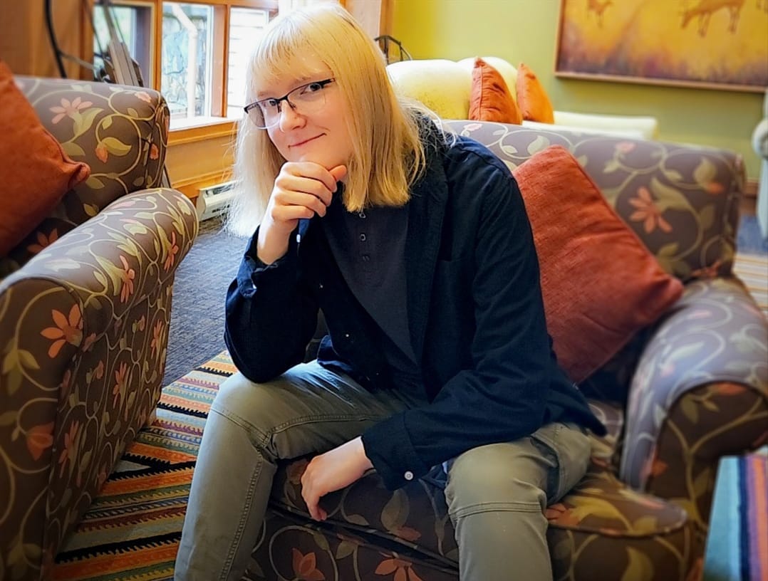 A person with light blonde hair and glasses sits on a floral-patterned armchair, resting their chin on their hand and smiling subtly. The image is found within the article: Scoring the Skies: Inside a CAT Grad's Journey from Student to Pro Composer.