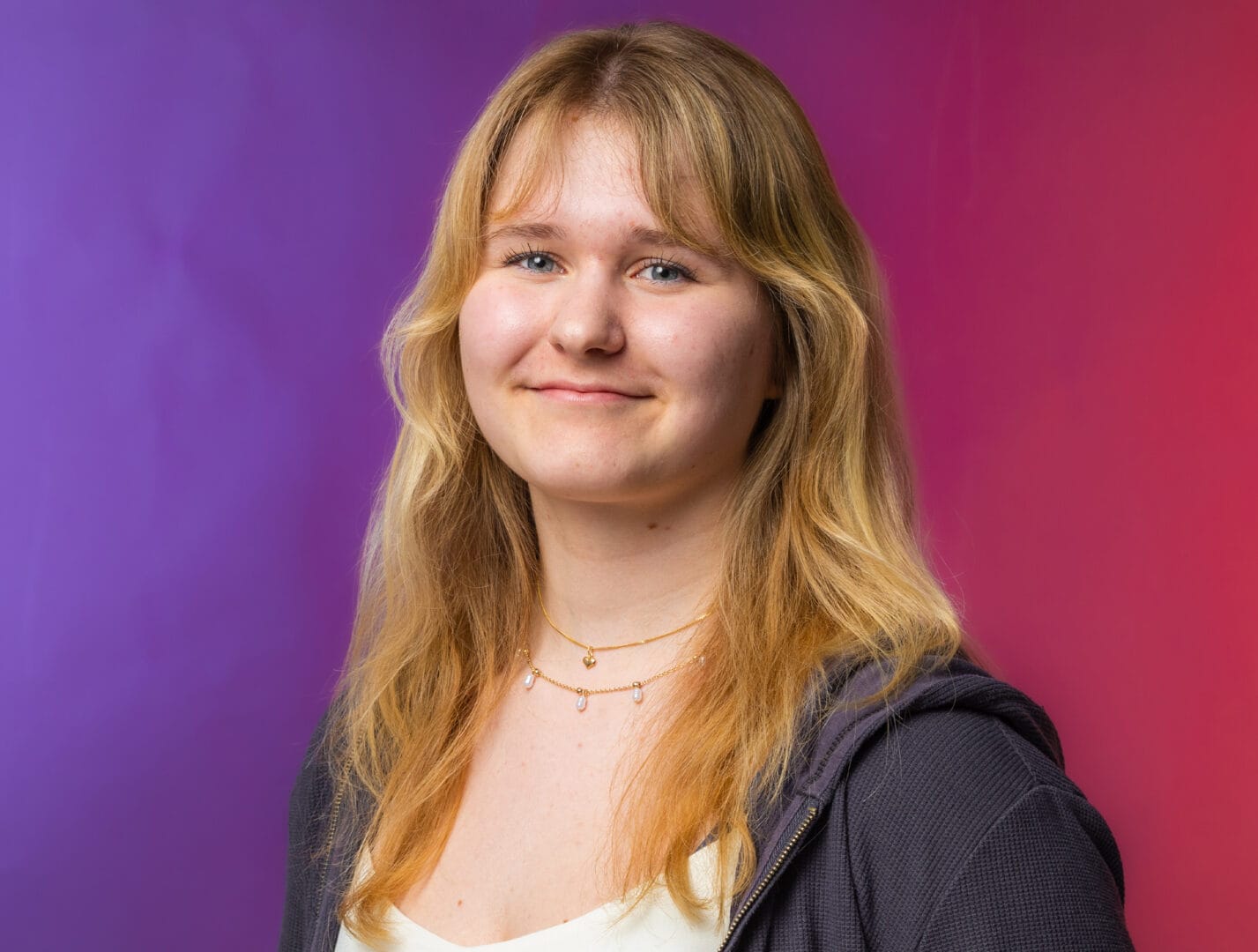 A young woman with long blonde hair, wearing a necklace and a dark jacket, stands against a gradient purple and red background.