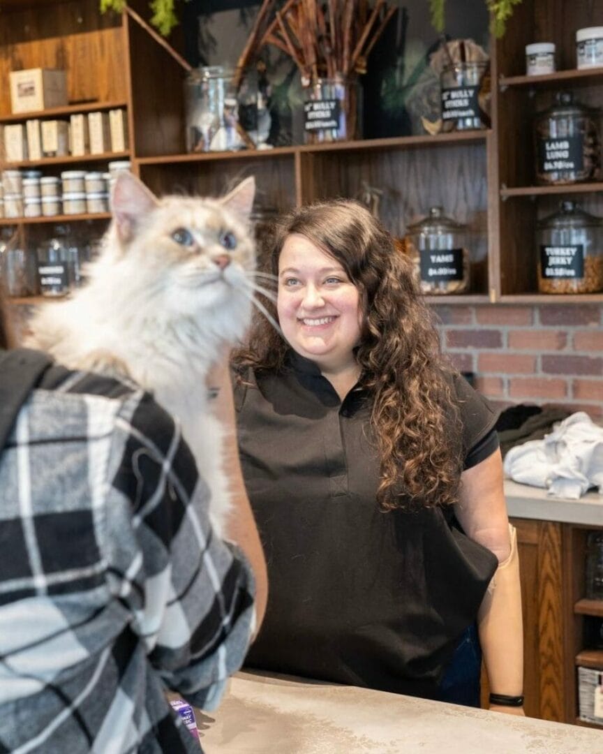 A smiling woman stands behind a counter in a store, looking at a fluffy cat perched on a person's shoulder in front of her. Shelves with jars and products are visible in the background.