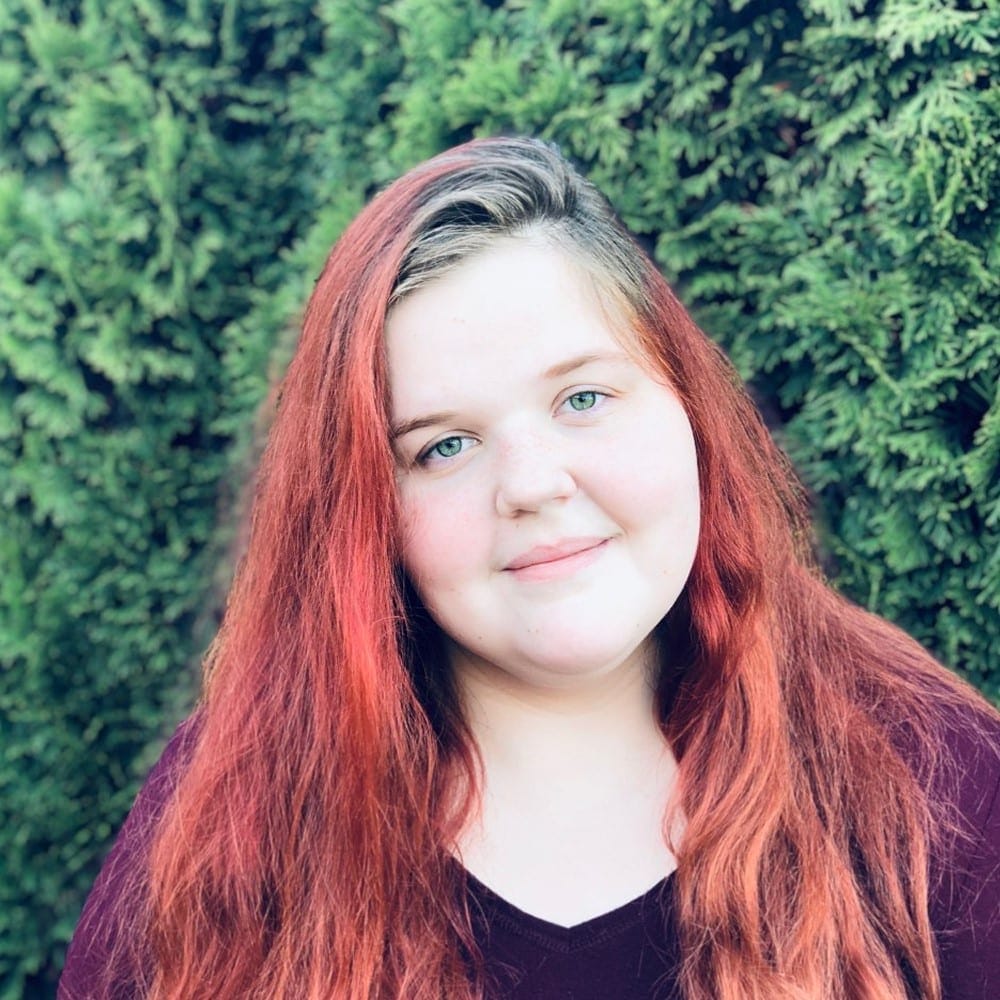 A girl with red hair standing in front of bushes.